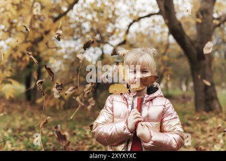 Bonne petite fille blonde joue avec des feuilles d'automne jaunes dans le jardin.L'enfant sourit et s'amuse au parc.Un gamin qui jette des feuilles en automne Banque D'Images