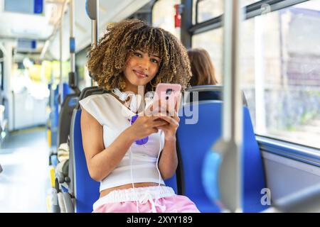 Jeune femme de beauté latine souriant à la caméra tout en utilisant le téléphone assis sur le bus urbain Banque D'Images