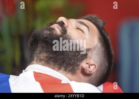 Barbier rasant la barbe d'un bel homme barbu avec un rasoir électrique au salon de coiffure Banque D'Images