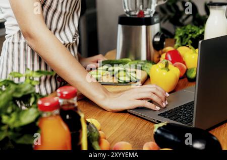Femme utilisant un ordinateur portable à la cuisine à la maison. Femme main clic, touchant l'ordinateur devant les légumes, les fruits sur la table à la maison. Mise au point sélective, recadrée Banque D'Images
