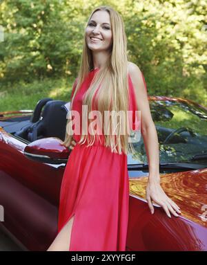 Belle jeune femme blonde en robe rouge debout près d'une voiture de sport Banque D'Images