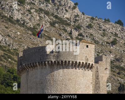 Gros plan d'une tour de château en pierre avec un drapeau flottant, entouré d'un paysage de montagne sous un ciel ensoleillé, Dubrovnik, mer Méditerranée, Croatie, UE Banque D'Images