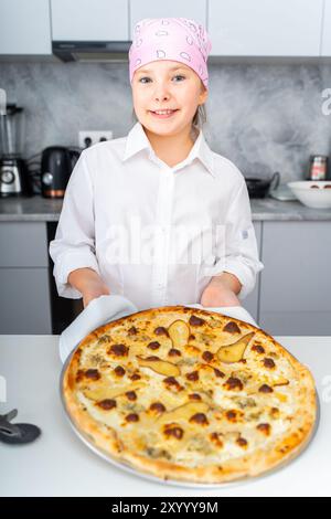 Une petite adolescente tient une pizza avec du fromage et de la poire, cuite pour elle à la maison dans la cuisine. Photo de haute qualité Banque D'Images