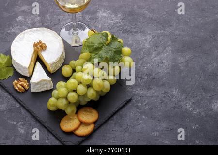 Planche d'ardoise avec brie de fromage, raisins blancs, craquelins noix et vin sur pierre noire, vue horizontale de dessus, espace de copie Banque D'Images