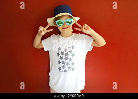 Très emothional enfant porter un chapeau et des lunettes de soleil sur fond rouge. Copie espace, lumière du jour Banque D'Images