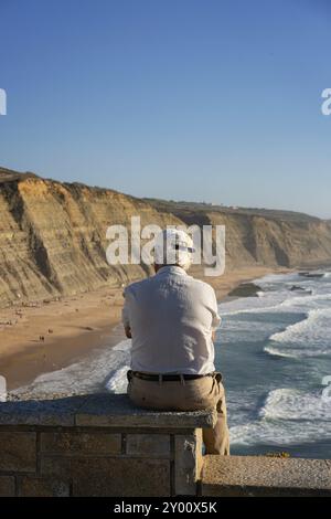 Vieil homme regardant la plage de Magoito avec des surfeurs surfant sur les vagues de la mer à Sintra, Portugal, Europe Banque D'Images