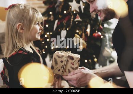 Un jeune père en chapeau de père Noël rouge donne un cadeau de Noël à sa fille souriante près d'un arbre de Noël décoré. Fille vêtue de Noël rouge-noir festif Banque D'Images