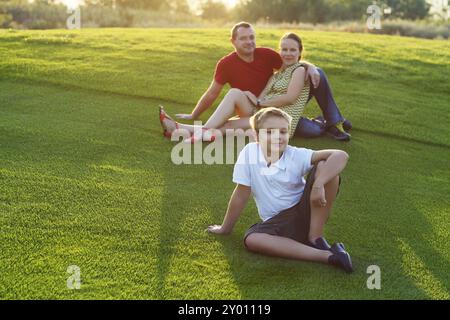 Famille heureuse avec fils assis sur l'herbe dans le parc. Coucher du soleil d'été Banque D'Images