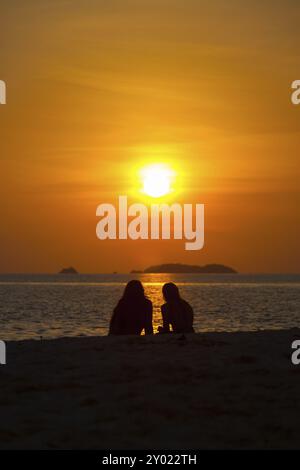 Deux personnes non identifiables, regarder un coucher de soleil dans l'océan sur une plage de sable de l'océan en face de ciel orange vif Banque D'Images