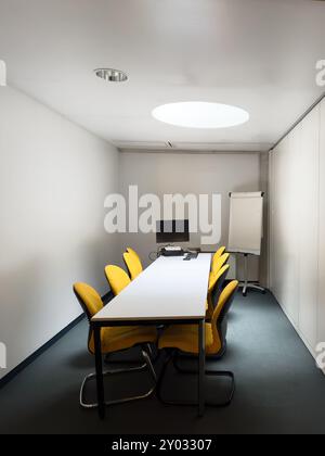 Une petite salle de conférence minimaliste avec des chaises jaunes autour d'une longue table, avec un ordinateur et un chevalet de conférence, prête pour une réunion ou une présentation dans un cadre d'entreprise. Banque D'Images