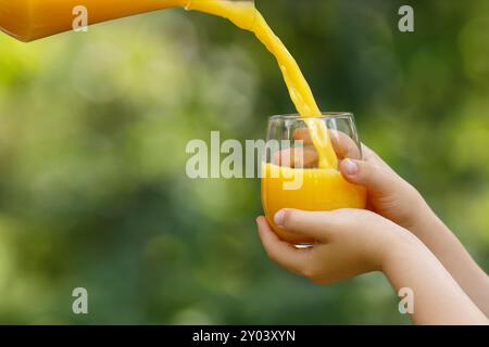 mains d'enfant tenant le verre et le jus d'orange coulant de la cruche avec jardin vert sur le fond Banque D'Images