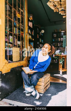 Femme souriante tenant le livre tout en étant assis sur la caisse près de la porte à la librairie Banque D'Images
