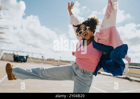 Joyeuse femme aux cheveux bouclés dans des lunettes de soleil et une veste letterman dansant sur le parking sous le ciel nuageux Banque D'Images