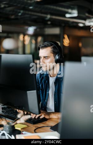 Jeune programmeur informatique tapant sur PC de bureau avec casque sans fil au bureau Banque D'Images