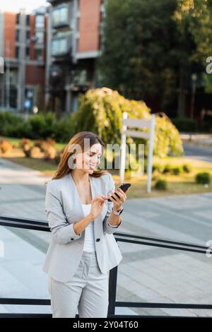 Une jeune femme confiante dans un costume gris clair s'engage avec son smartphone, rayonnant de positivité alors qu'elle apprécie l'atmosphère urbaine animée remplie de gre Banque D'Images