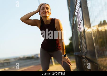 Femme fait une pause pendant son entraînement du soir, essuyant son front alors que le soleil se couche, entourée d'un chemin de jogging tranquille et d'un paysage légèrement éclairé Banque D'Images