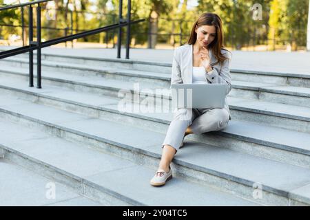 Jeune femme d'affaires dans une tenue chic s'assoit confortablement sur les marches, engrossée dans son ordinateur portable, comme la lumière chaude du soleil projette des ombres douces, mélangeant produc Banque D'Images