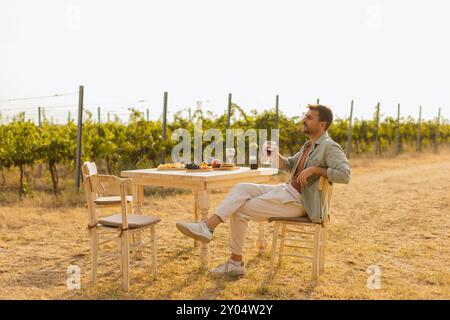 L'homme profite d'un moment de paix à une table rustique au milieu des rangées de vignes luxuriantes, en savourant du vin et des fruits frais tout en trempant dans la lumière dorée de l'après-midi Banque D'Images