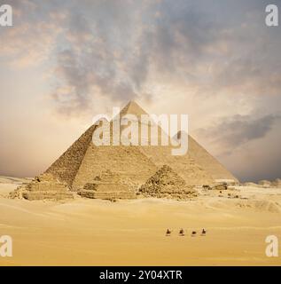 Coucher de soleil spectaculaire derrière toutes les pyramides égyptiennes lointaines avec une ligne de chameaux marchant au premier plan à Gizeh, le Caire, Egypte. Téléphone compressé large Banque D'Images