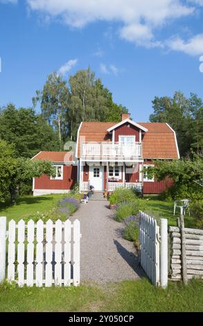 Maison suédoise traditionnelle en bois peint en rouge dans un jardin verdoyant avec une clôture blanche de piquet et une porte ouverte avec une passerelle menant à la porte d'entrée Banque D'Images