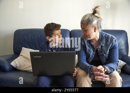 Mère et fils sont assis sur un canapé bleu, engagés dans la conversation tout en regardant un ordinateur portable, dans le salon de leur maison Banque D'Images