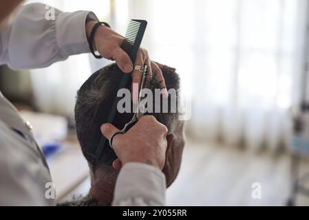 Un coiffeur en chemise blanche coupe les cheveux d'un homme avec une paire de ciseaux et un peigne à la maison Banque D'Images