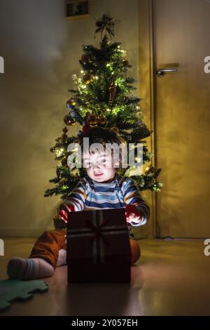 Un jeune enfant est assis devant un sapin de Noël, ouvrant un cadeau rouge. La scène est chaleureuse et festive, avec l'excitation de l'enfant et la déco de l'arbre Banque D'Images