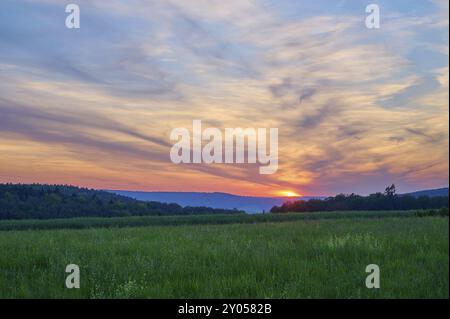 Champ vert avec des arbres en arrière-plan sous un coucher de soleil dramatiquement illuminé, le ciel est baigné de tons orange et bleu, été, Moenchberg, Mlte Banque D'Images