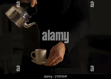 Les mains d'un homme méconnaissable dans l'ombre versant une tasse de café d'une théière en métal Banque D'Images