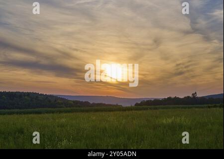 Champ vert avec des arbres en arrière-plan sous un coucher de soleil dramatiquement illuminé, le ciel est baigné de tons orange et bleu, Moenchberg, Mltenberg, S. Banque D'Images