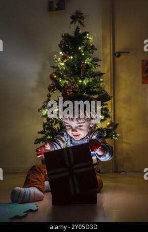 Un jeune garçon ouvre une boîte rouge avec un arc blanc dessus. Il est assis par terre devant un sapin de Noël Banque D'Images