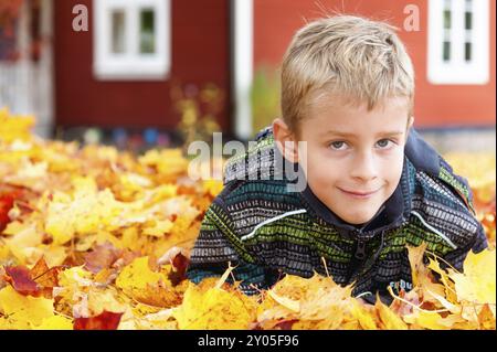 Mignon petit garçon joue dans les feuilles d'automne et sourit malicieusement dans la caméra. En arrière-plan, une maison en bois typiquement suédoise peinte en rouge. Mignon y Banque D'Images