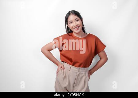Belle jeune femme asiatique en chemise brune et pantalon taupe plaçant les mains sur la hanche avec une expression confiante, isolée par fond blanc. Banque D'Images
