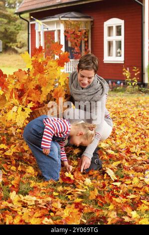 Riant jeune mère et sa jolie petite fille jouant dans des feuilles d'automne colorées jaune et orange tout en collectant un basket de brindilles feuillues à Banque D'Images