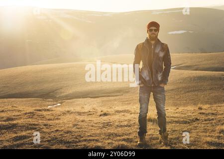 Portrait de mode d'un jeune homme hipster barbu portant des lunettes de soleil, un sac à dos et un chapeau sur un fond avec copyspase dans les montagnes au coucher du soleil, Un co Banque D'Images