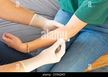 Docteur en vêtements de plaine traitant le bras d'une femme. Soins à domicile pour une fille. Médecin sans manteau blanc avec des gants blancs donnant des soins médicaux à domicile. Banque D'Images