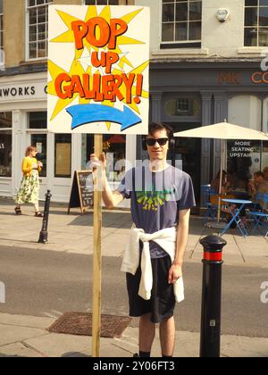 Homme portant des lunettes de soleil et des vêtements d'été tenant des panneaux publicitaires aux couleurs vives Galerie pop up dans le centre-ville de Bath. Banque D'Images