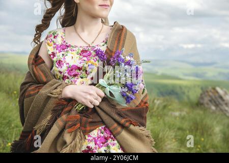 Gros plan d'une fille rustique. Une fille enveloppée dans un plaid et dans une robe colorée tient un bouquet de fleurs sauvages Banque D'Images