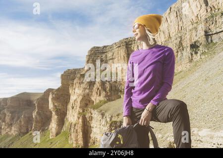 Voyageuse fille souriante dans un chapeau jaune et une paire de lunettes de soleil se tient au pied de rochers épiques avec un sac à dos à côté et regarde loin Banque D'Images