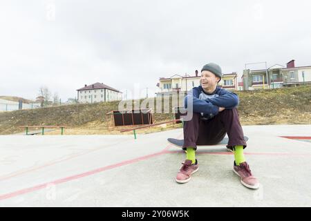 Un adolescent est assis sur un skateboard dans le parc et souriant. Le concept de temps libre passe pour les adolescents dans la ville Banque D'Images