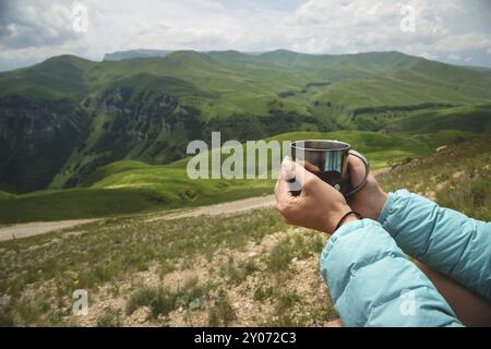 Vue de la première personne des mains d'une fille tenant une tasse de thé en plastique contre un plateau de collines vertes et un ciel nuageux en été Banque D'Images