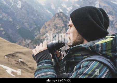 Un homme hipster avec une barbe dans un chapeau, une veste et un sac à dos dans les montagnes caucasiennes tient des jumelles, aventure, tourisme, pistage Banque D'Images