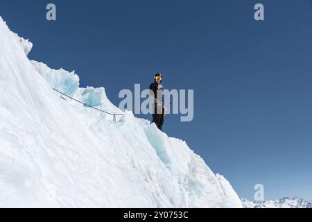 Un alpiniste se tient au bord d'un glacier avec une pelle à neige entre ses mains et montre le geste de Shak contre le ciel bleu Banque D'Images