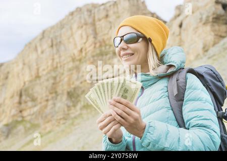 Un voyageur dans un chapeau et des lunettes de soleil tient une centaine de billets de dollars dans les mains d'un ventilateur sur fond de rochers sur la nature. Frais de déplacement Banque D'Images