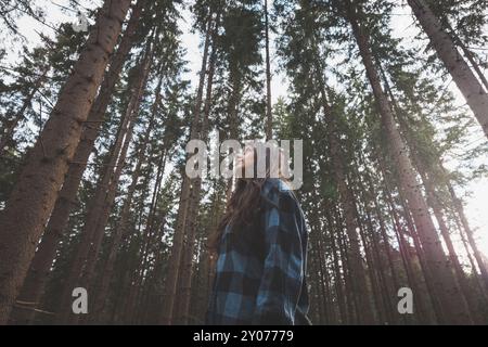 Femme heureuse dans une chemise à carreaux profitant de la sérénité d'une forêt dense, entourée de grands arbres, se sentant libre et en connexion avec la nature. Banque D'Images