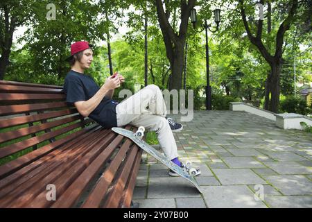 Un patineur hipster aux cheveux longs dans une casquette est assis sur un banc et tourne une spineless spinner à côté d'une planche à roulettes Banque D'Images