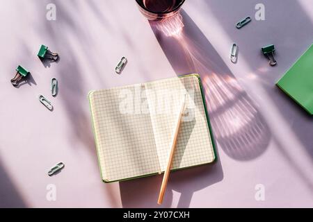 Papeterie d'été moderne nature morte. Un verre d'eau avec une longue ombre et un bloc-notes ouvert avec un crayon sur fond violet. Vue de dessus, pose à plat. Banque D'Images