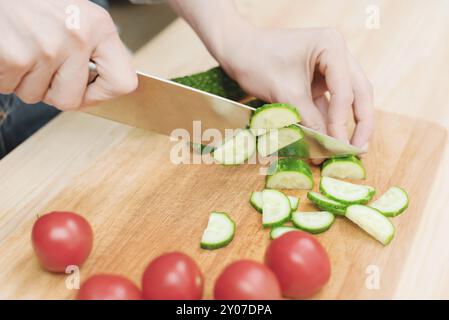 Gros plan des mains femelles coupées en concombres frais coupés sur une planche à découper en bois à côté des tomates roses. Le concept de cuisine végétarienne maison et Banque D'Images