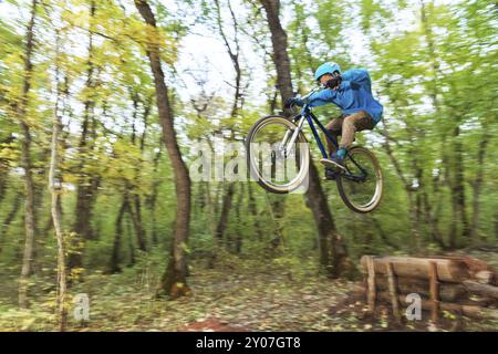 Un jeune cavalier dans un casque et un sweat-shirt bleu vole sur un vélo après avoir sauté d'un haut kicker sur une piste cyclable forestière. Prise de vue avec exposition prolongée Banque D'Images