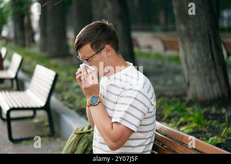 Homme dans la chemise rayée éternue ou souffle le nez dans la serviette tout en étant assis sur le banc dans le parc de la ville. Allergies saisonnières à la floraison ou au rhume, période de floraison et propagation des peluches et du pollen à l'extérieur, mauvaise écologie Banque D'Images